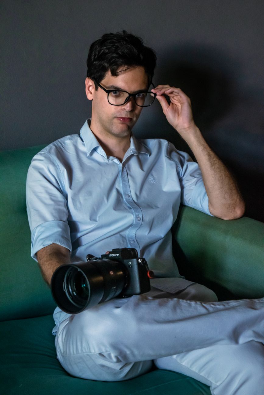 Young male photographer holding a camera, sitting on a couch indoors.